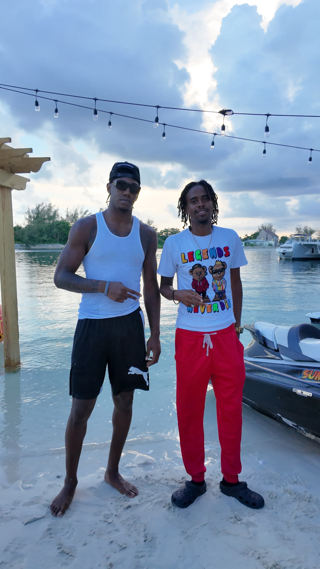 Two men posing on a beach at dusk beside a jet ski under string lights