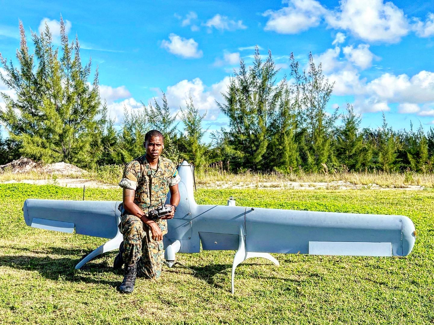FlyWright pilot in uniform kneeling beside a fixed-wing unmanned aircraft in an open field, radio controller in hand