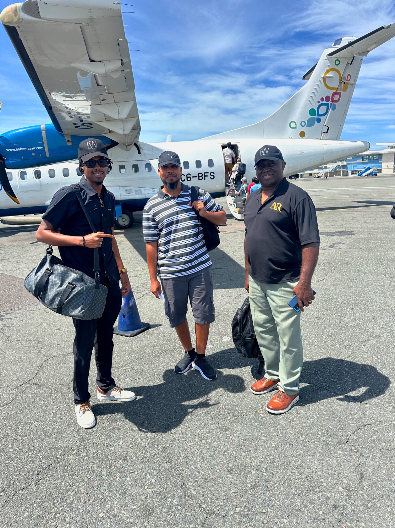 Three men in FlyWright caps standing on an airstrip in front of a Bahamasair aircraft